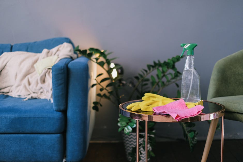 A person wearing a plaid shirt and beige trousers is using a fluffy, blue dusting tool to clean the top of a brown fabric sofa in a bright living room. The sofa has an orange cushion resting against i