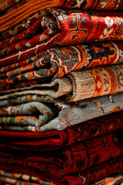 Close-up view of several hanging rugs with intricate, colorful patterns including geometric shapes and floral designs, displayed vertically in a well-lit indoor setting. The rugs feature a variety of